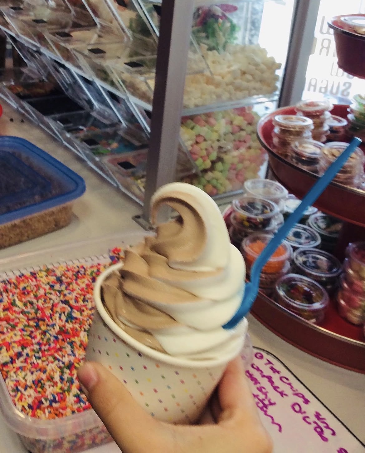 Vanilla and chocolate soft serve swirl in a cup, with toppings and candy in the background